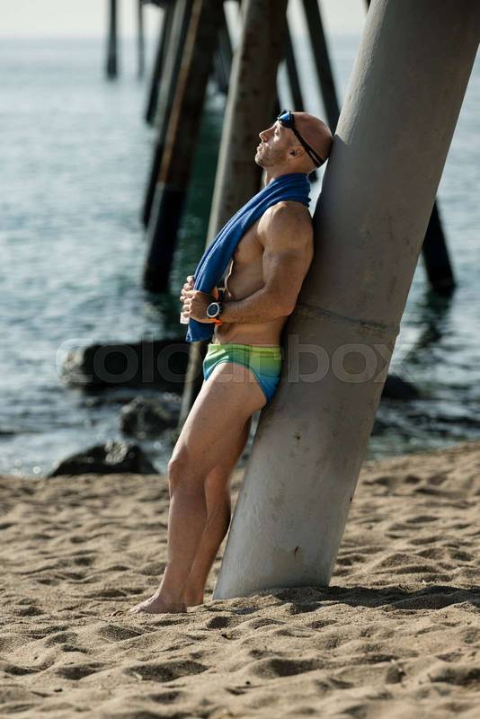 Professional swimmer toweling off his head with swim glasses sitting at the beach. Fit man with a blue towel after training leaning against stones wearing speedo.