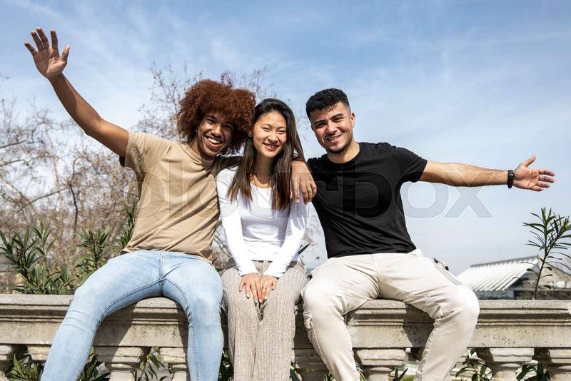Multiracial group of young smiling people sitting on a stone balcony. Three diverse happy friends having fun together in a park.
