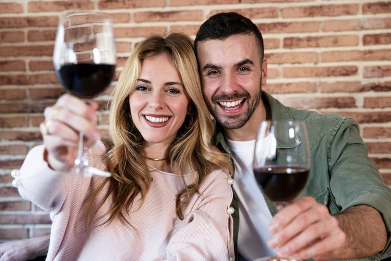  Cheerful male and female drinking with glasses at home. Joyful young adult couple toasting with wine and looking at camera.