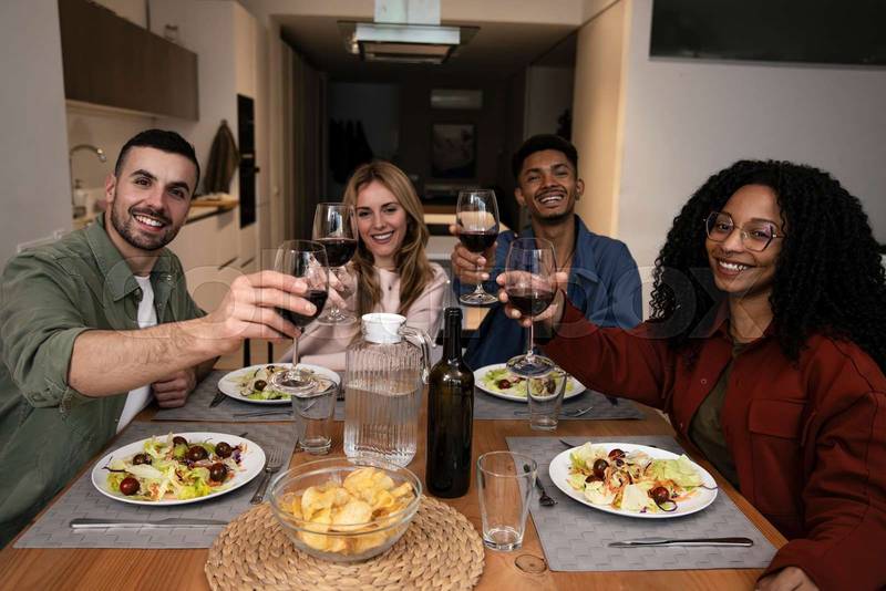 Smiling people toasting eating and drinking on a modern apartment. Diverse group of happy friends celebrating diner at home looking at camera. 