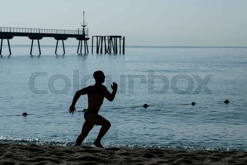 Running man silhouette training at the beach. Athlete jogging next to the sea with a sea dock in background.