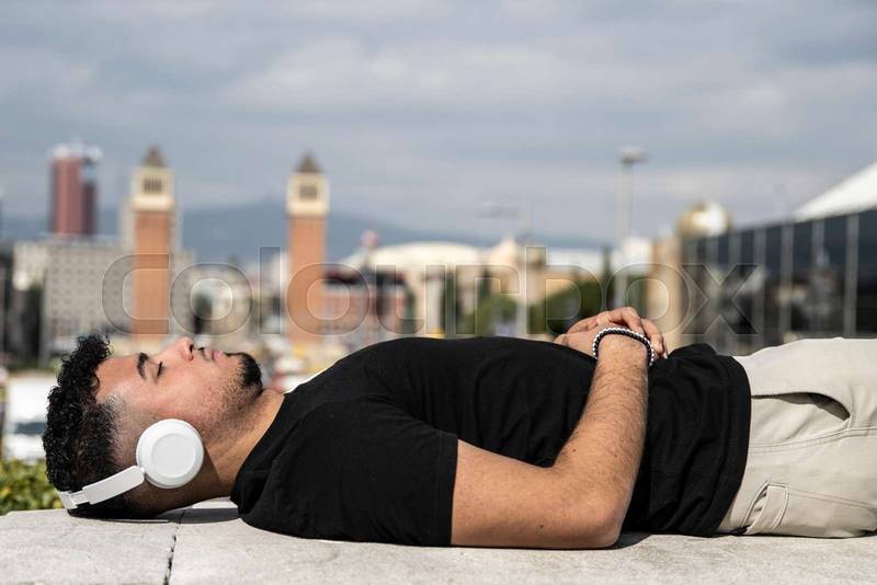 Relaxed young man lying in a balcony while listening music with headphones. Confident young guy lying down with closed eyes listening to the radio with the city landscape in the background.