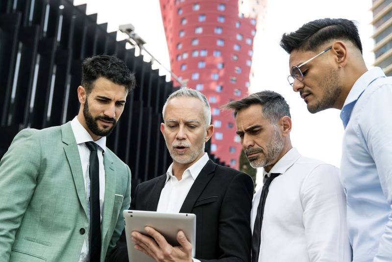 Diverse group of businessman checking a tablet in the street. Senior ceo holding a device in a meeting with young colleagues outside of office buildings.