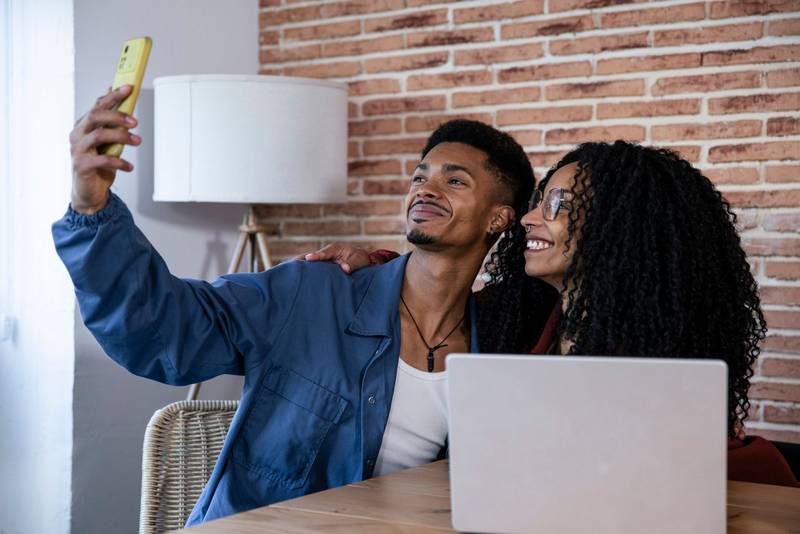 Young couple taking selfie while using computer at home. Young adult friends with laptop and mobile phone taking a picture while sitting on the living room.