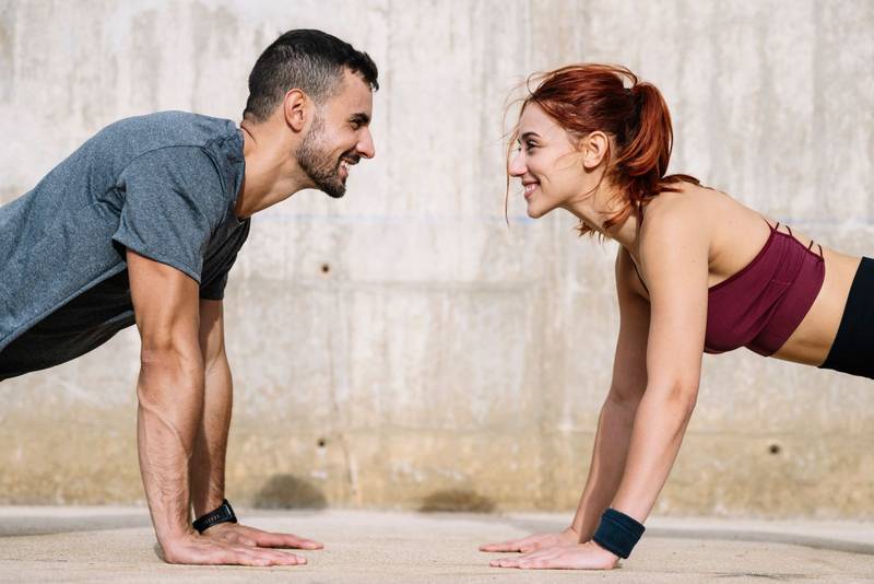 Determined young adult couple exercising in the floor looking each other. Joyful athletic male and female doing push ups in street in sportswear training workout outside. 