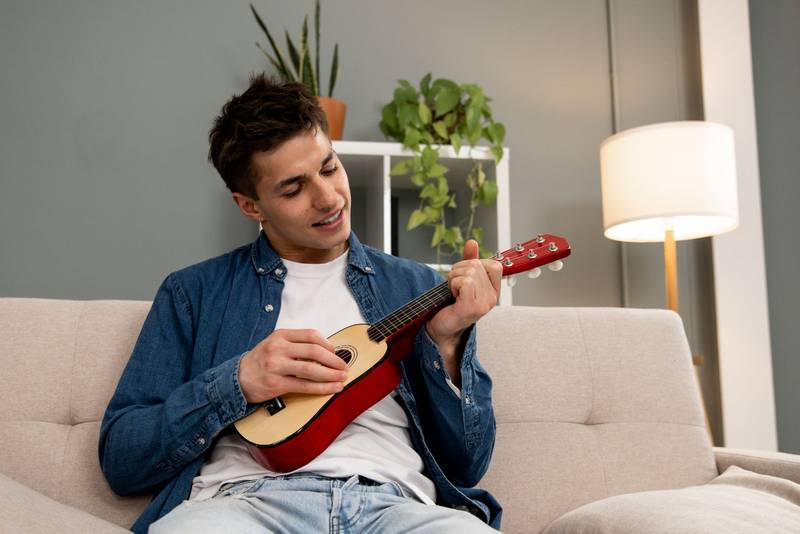 Young relaxed man playing music with ukelele sitting on sofa at home. Young adult carefree male learning to play guitar on couch at the living room.