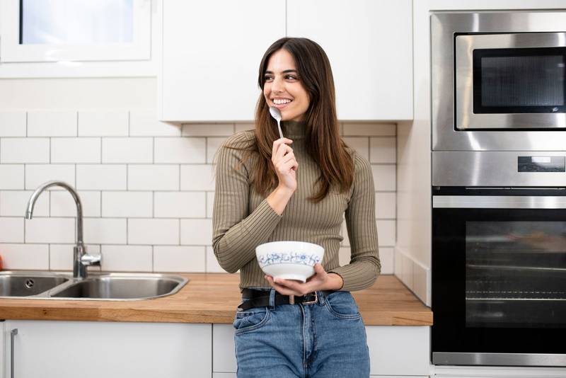 Portrait of beautiful young woman having breakfast in the kitchen. smiling female with a bowl ans spoon looking at camera. Lifestyle concept.