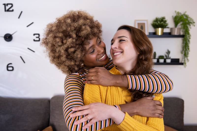 Beautiful and cheerful multiracial lesbian couple hugging and looking each other smiling. Two lovely and happy homosexual women embracing at home staring each other.