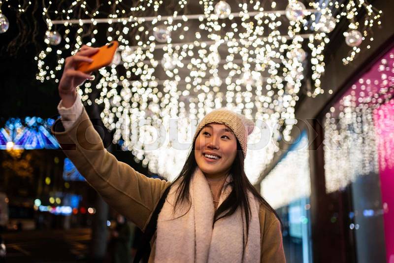 Young cheerful girl taking a selfie in the street at christmas night. Happy woman wearing warm hat taking picture with her phone in winter.