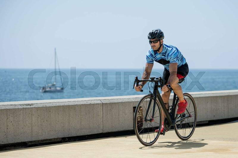 Athletic biker riding a bike in the street next to the sea. Fit cyclist training wearing helmet and sunglasses outdoors.