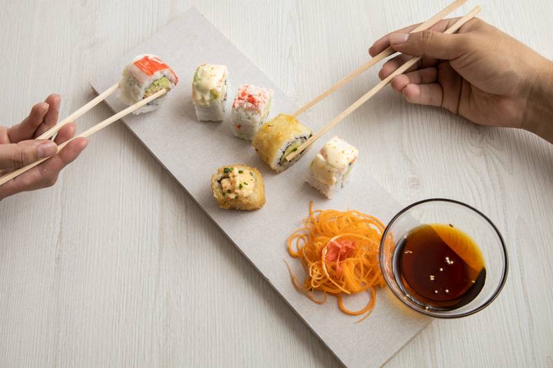 various sushi rolls of different flavors, carrot, soy sauce on a plate while two persons hand hold with chopsticks