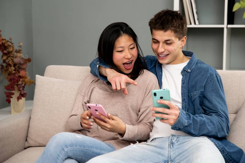 Affectionate young couple with mobile phones relaxing on sofa. Happy diverse woman and man hugging using smartphone together sitting on cozy couch at home.