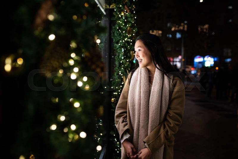 Hopeful young girl looking at shop window in a winter night. Smiling female holding bags with Christmas decoration shop.