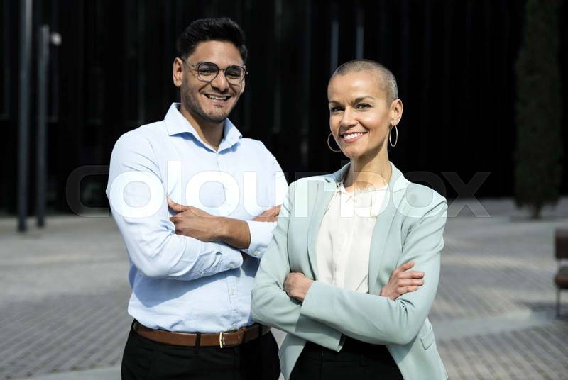 Happy multicultural business couple posing with arms crossed in outdoors. Portrait of multiethnic corporate partenrs looking at camera. Finance concept.