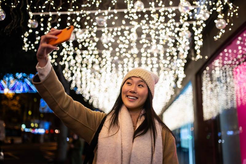 Young cheerful girl taking a selfie in the street at christmas night. Happy woman wearing warm hat taking picture with her phone in winter.