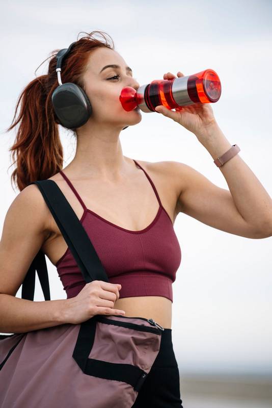 Young fitness female drinking water outside wearing headphones. Athletic woman using a bottle and a gym bag ready for training and workout.