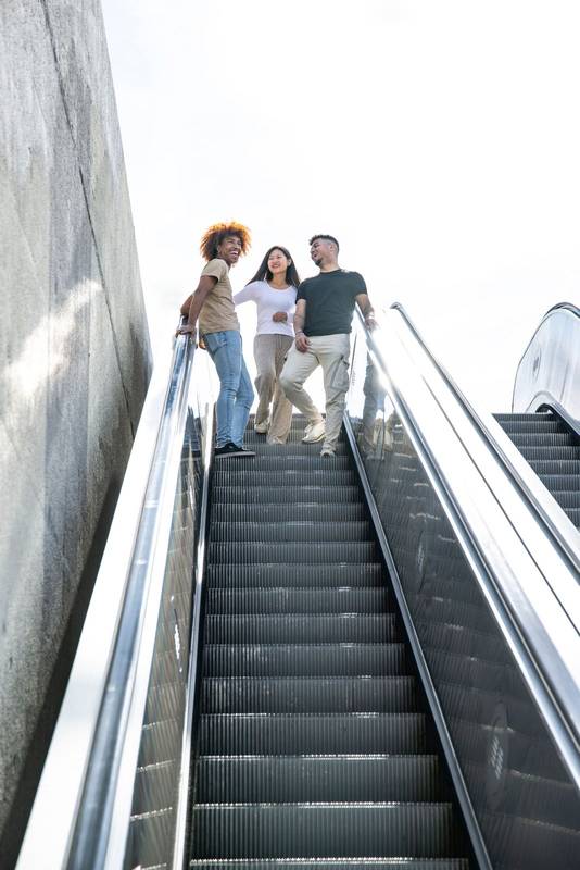Multiethnic group of friends laughing and going down an outdoor escalator. Three joyful young people smiling and looking relaxed walking down from the top of a stairs in the street.