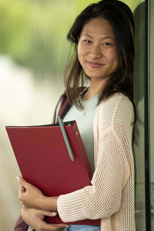 Handsome satisfied student holding a notebook looking at camera leaning against a glass wall.Confident and happy woman standing outside with files on her hands.