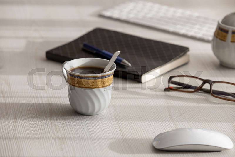 desk with a cup of coffee with a spoon, it is next to glasses, computer mouse, sugar bowl, notebook, pen and keyboard
