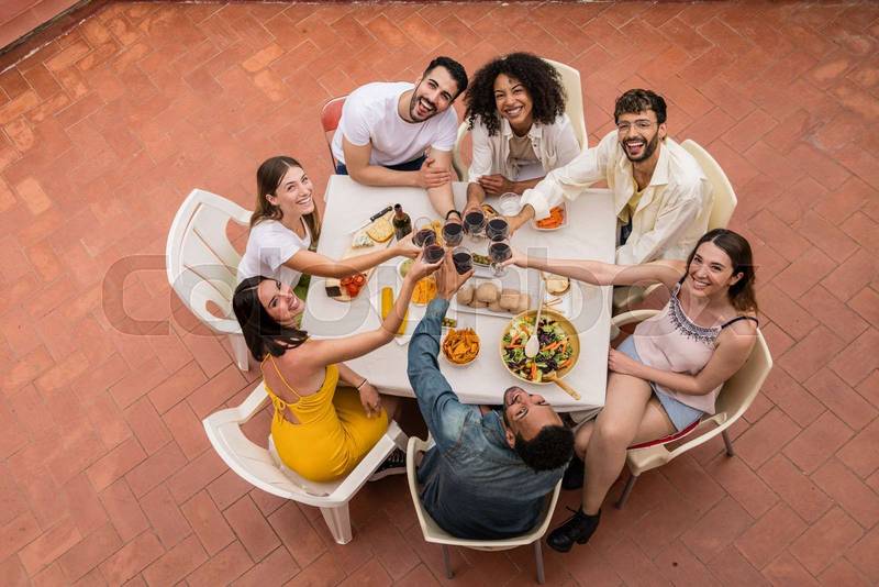 Top view of a group of happy young friends toasting with glasses in a terrace. Carefree and cheerful people sitting in a patio having dinner and drinking wine.