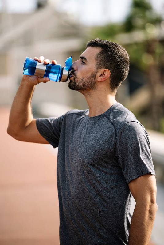 Young sports man drinking water from a bottle resting after workout in a park. Healthy fit male drinks and recovery with refreshment after exercise outside.