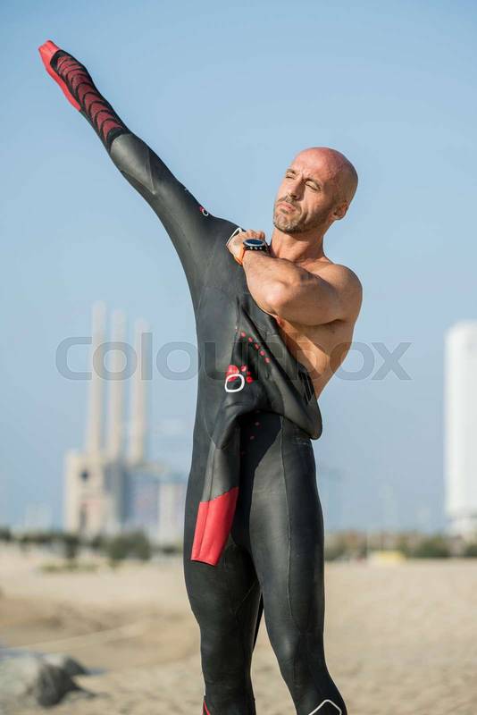 Male swimmer getting ready for swim training in the beach with urban background. Confident man putting on a wetsuit to go swimming in the ocean.