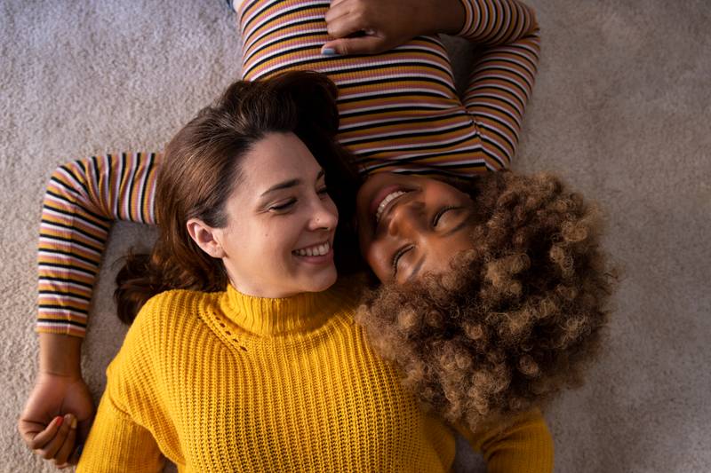 Top view of lovely multiracial lesbian couple lying on a carpet looking aat each other. Two beautiful happy homosexual women smiling and relaxing together on the floor. Relationship concept.