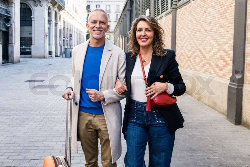 Affectionate senior man and woman standing and holding arms during a touristic visit to an european city. Mature couple bonding together in a romantic trip looking at camera and holding suitcase.