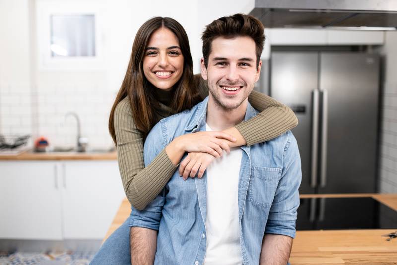 portrait of smiling young couple in the kitchen at home. Handsome man and beautiful woman standing together ans looking at camera. Lifestyle concept.