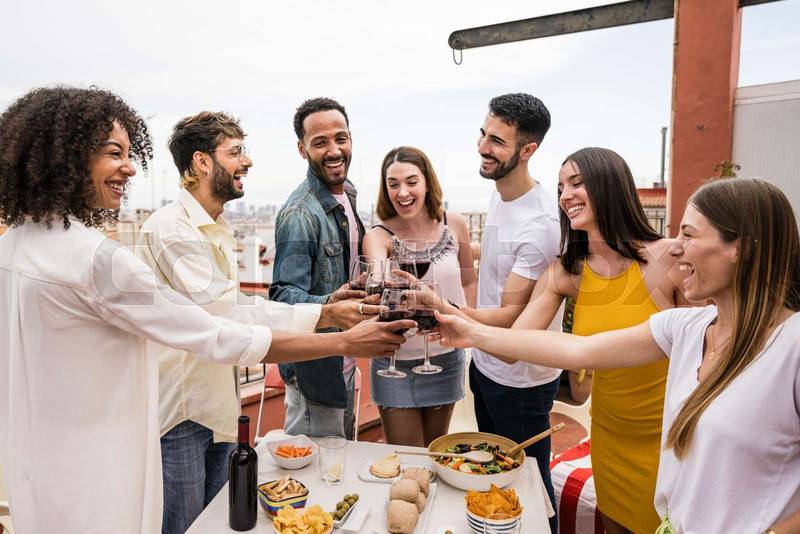 Happy group of friends toasting with wine glasses standing around a table in a terrace. Diverse group of cheerful young people drinking and celebrating in a rooftop dinner.