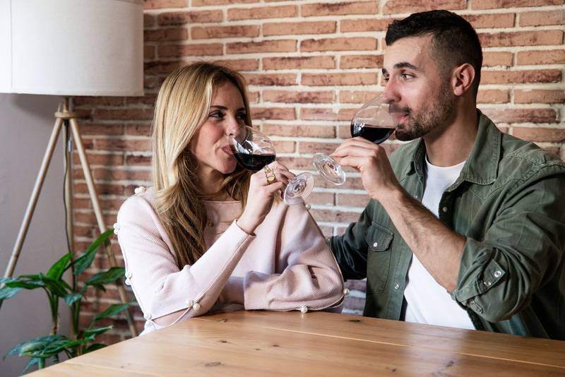 Happy woman and man drinking wine together in a living room with glasses. Lovely couple smiling in a date drinking alcohol sitting at table in a house.