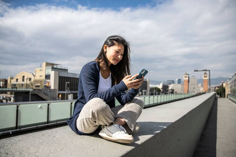 Young beautiful girl sitting on bank relaxed and texting with her phone. Satisfied woman using an smartphone sitting in the street.