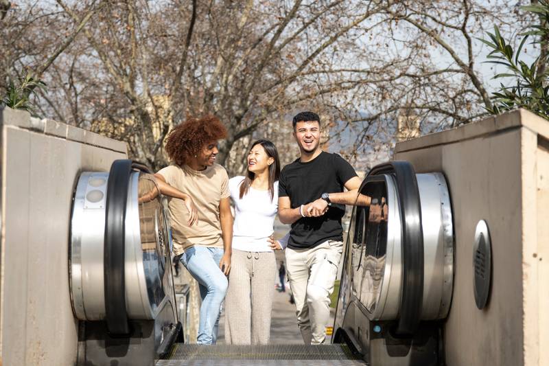 Diverse group of friends going up an outdoor escalator in a park. Three happy young people smiling and looking relaxed climbing up the stairs in the street.