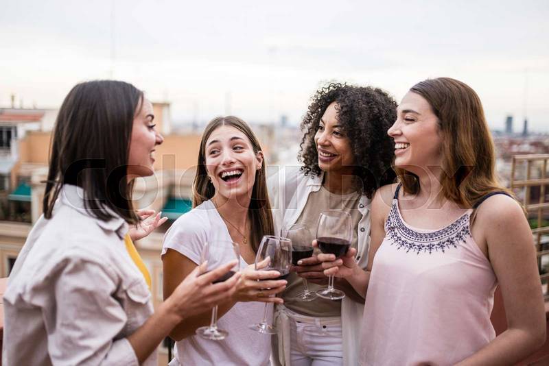 Diverse group of female friends having fun while drinking wine in a rooftop party. Carefree and happy young women laughing standing in a terrace holding glasses.