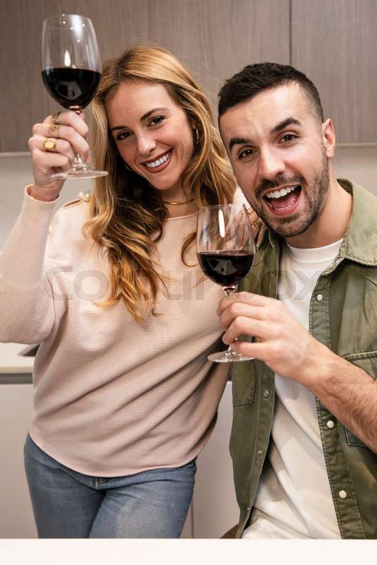Affectionate joyful couple celebrating toasting with wine in kitchen. Happy smiling man and woman drinking with glasses in home.