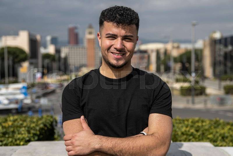 Young handsome guy smiling relaxed and looking at camera in the street with arms crossed. Confident happy man relaxed and staring at camera standing with arms folded outdoors.