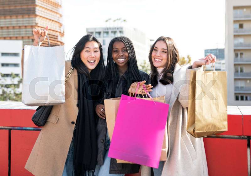 Three cheerful woman holding shopping bags in hand - Three girls, multiracial group, having fun in the city while shopping.