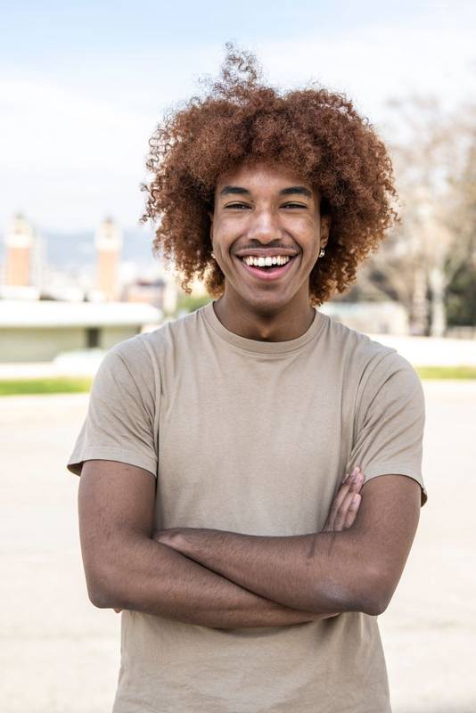 Young handsome guy smiling relaxed and looking at camera in the street with arms crossed. Confident happy man laughing and staring at camera standing with arms folded outdoors.