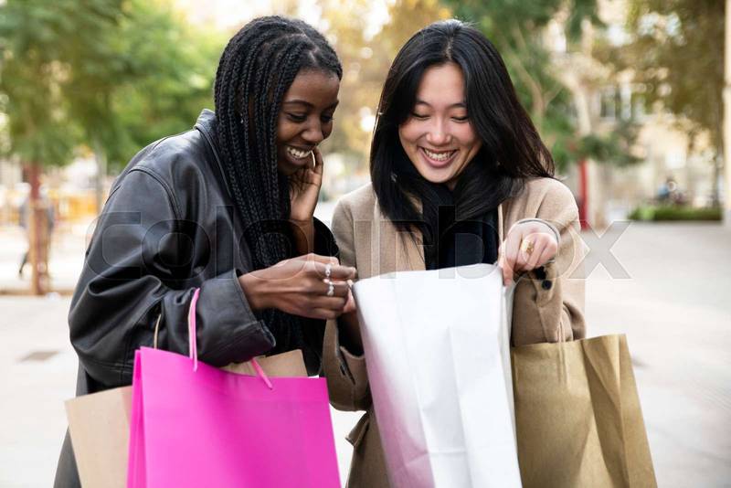 Happy beautiful women looking inside shopping bag and smiling at city outdoors . Fashion girl showing something to her friend in a shopping day