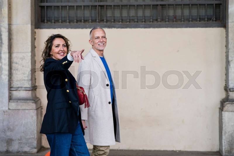 Affectionate mid adult tourist couple walking together pointing to some location. Lovely happy senior man and woman traveling and visiting streets of an old town.