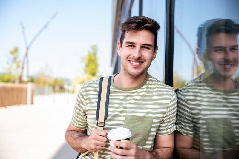 Happy young student smiling with backpack drinking a take away coffee at the campus.