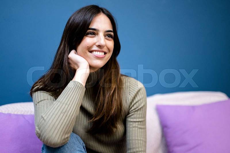 Happy young woman sitting on sofa at home and looking at camera. Portrait of comfortable caucasian woman similing and relaxing on armchair
