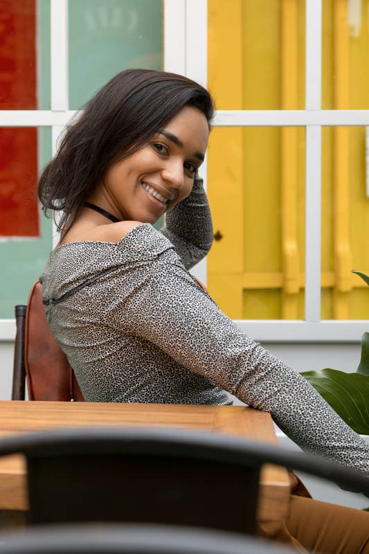 Inside a colorful house a young brunette woman with short hair smiling and sitting at a wooden table posing