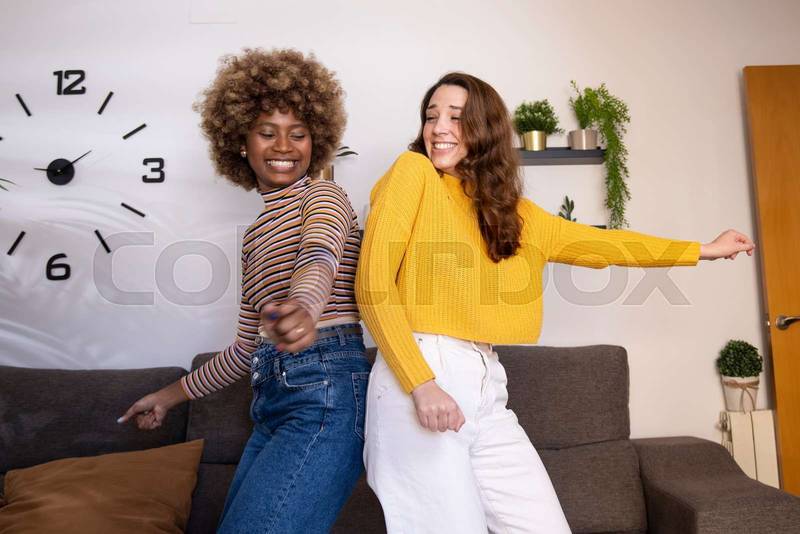 Multiracial young funny women dancing and laughing back to back in their apartment. Side view of happy and excited couple of girls celebrating in their living room.