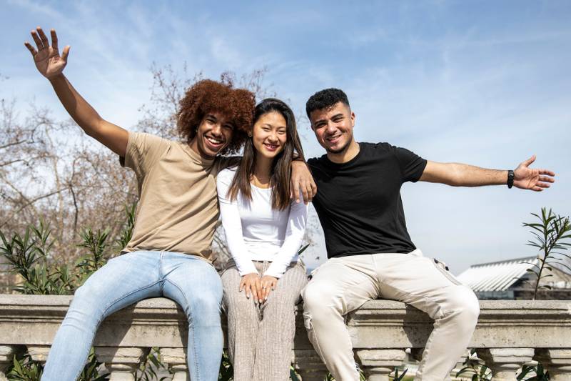 Multiracial group of young smiling people sitting on a stone balcony. Three diverse happy friends having fun together in a park.