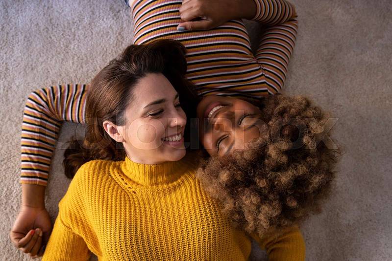 Top view of lovely multiracial lesbian couple lying on a carpet looking aat each other. Two beautiful happy homosexual women smiling and relaxing together on the floor. Relationship concept.