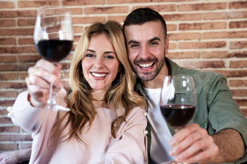  Cheerful male and female drinking with glasses at home. Joyful young adult couple toasting with wine and looking at camera.