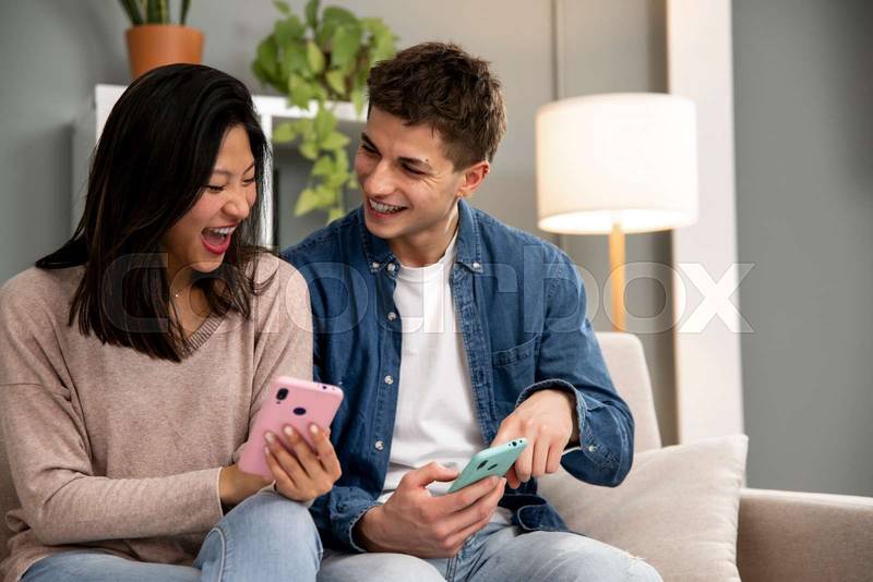 Happy young couple using smartphone relaxing on a couch in their living room at home. Diverse man and woman smiling and having fun using online phones.