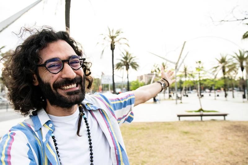 Portrait of cheerful happy hipster young man smiling looking at camera taking a selfie in the street with one arm wide open. Joyful carefree young guy laughing and staring at camera in the city.