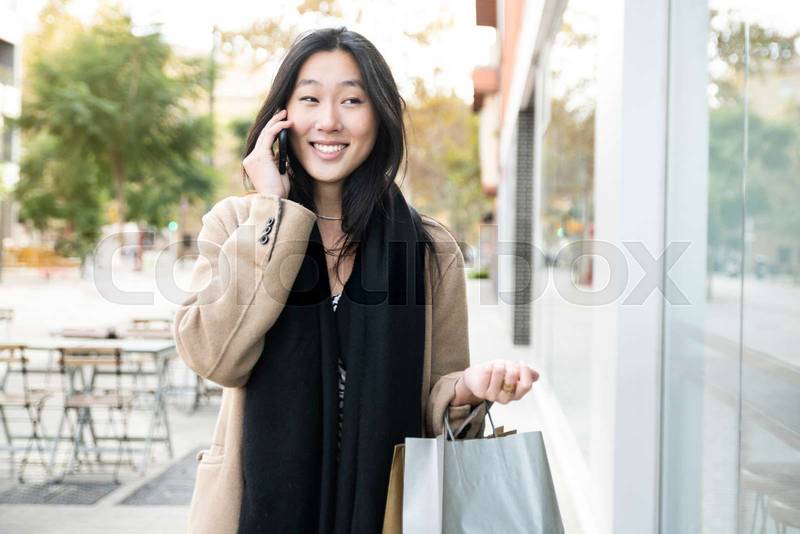 smiling young asian woman talking on a phone and holding shopping bags. happy female with a cellular in the city. Beauty, fashion, concept